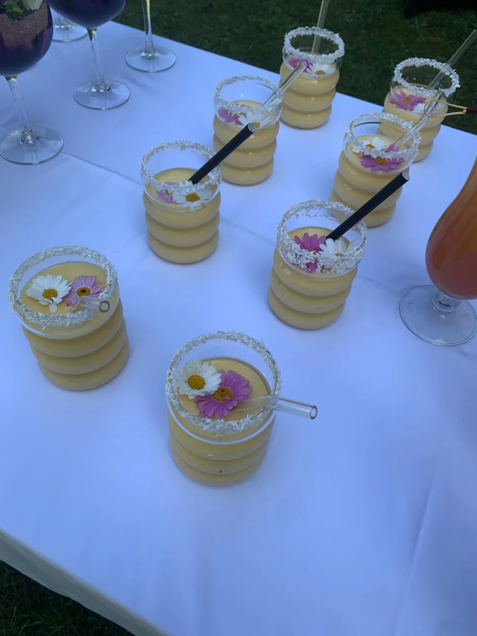 Petits verres de crème à étages décorés de fleurs comestibles, posés sur une table de réception en extérieur