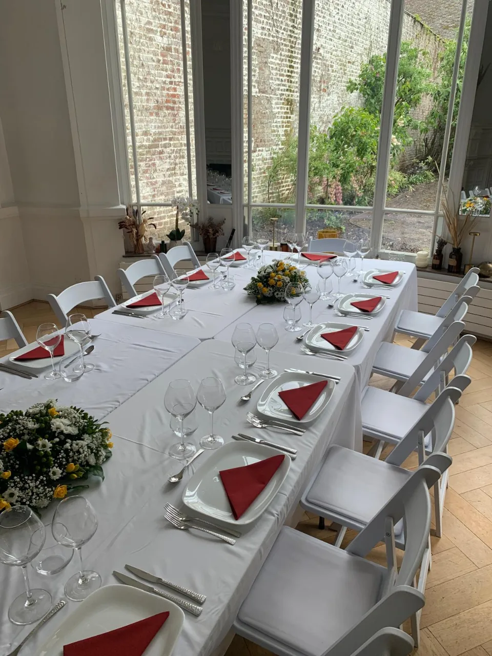 Longue table nappée de blanc avec serviettes rouges et centres de table fleuris, face à de grandes fenêtres