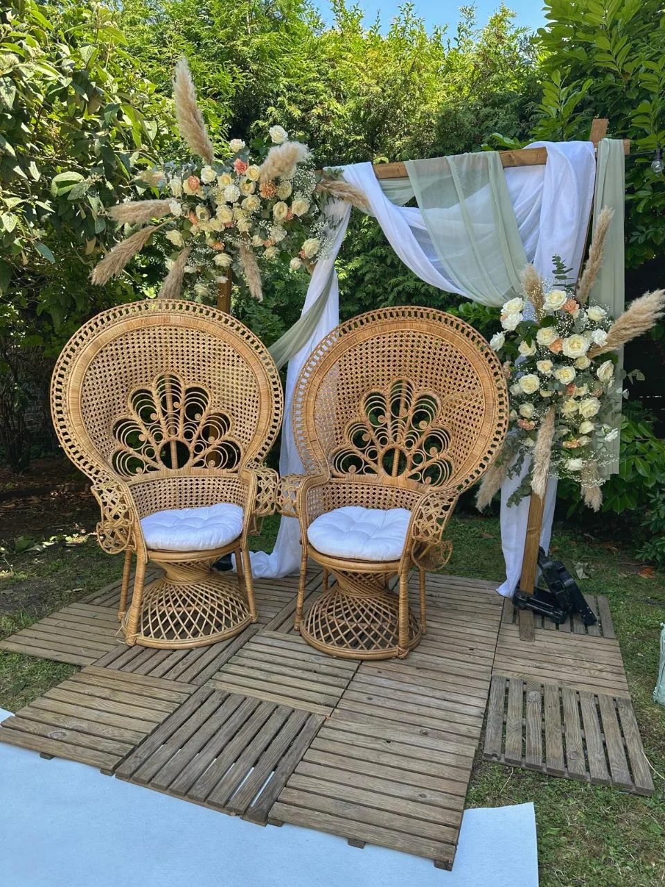 Deux fauteuils en rotin en forme de paon sous une arche décorée de tissu blanc et de fleurs, pour une cérémonie laïque