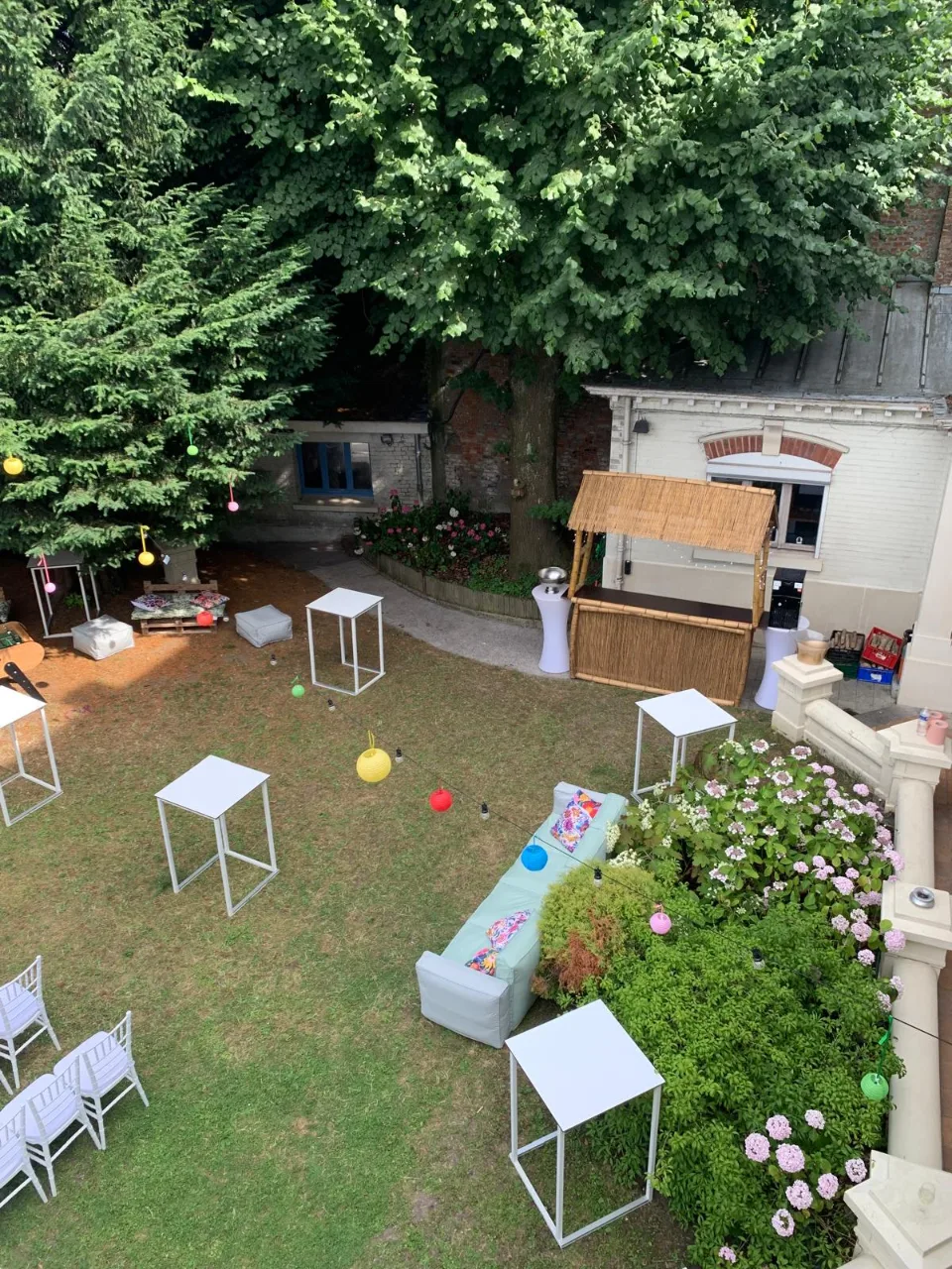 Vue du jardin avec tables hautes blanches, canapé de jardin fleuri et buvette en bois
