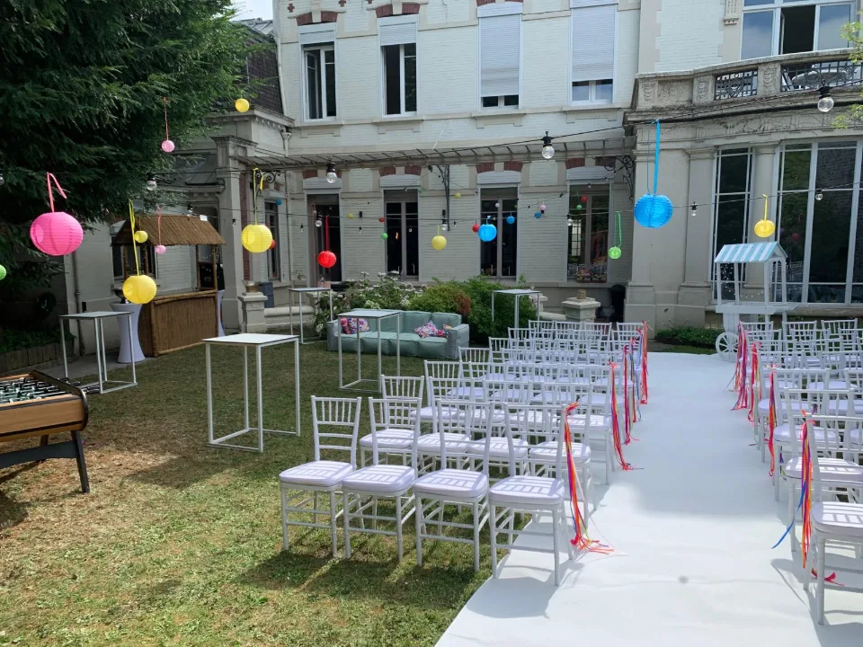 Chaises blanches alignées de chaque côté d'un tapis blanc, lampions colorés et façade de la maison au fond