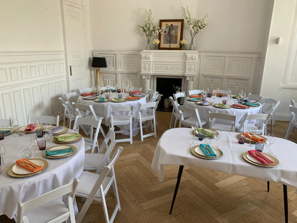 Salle avec cheminée et tables dressées d'assiettes colorées, nappes blanches et chaises pliantes