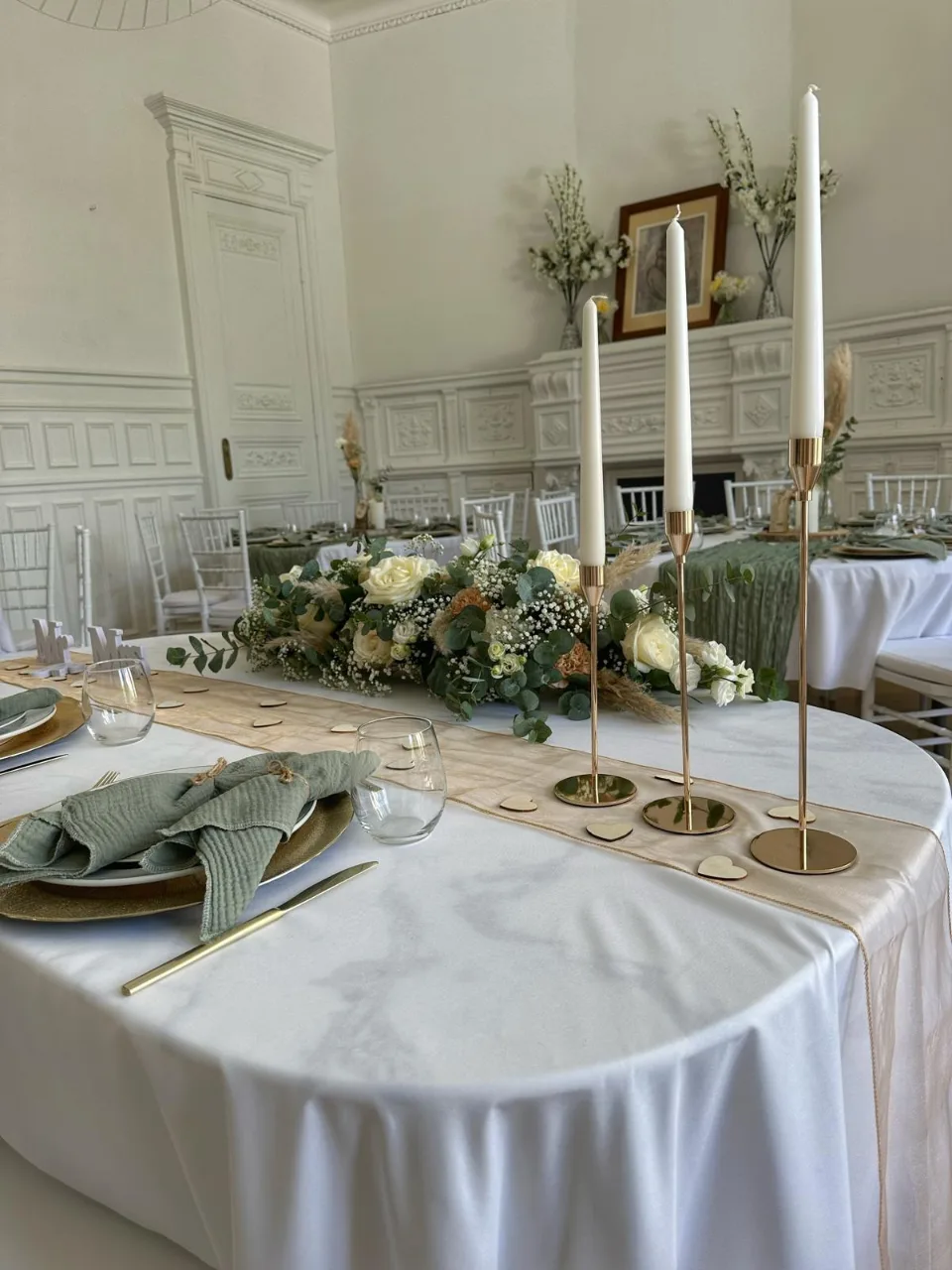 Table avec chandeliers dorés, composition florale et serviette vert sauge dans la salle de réception